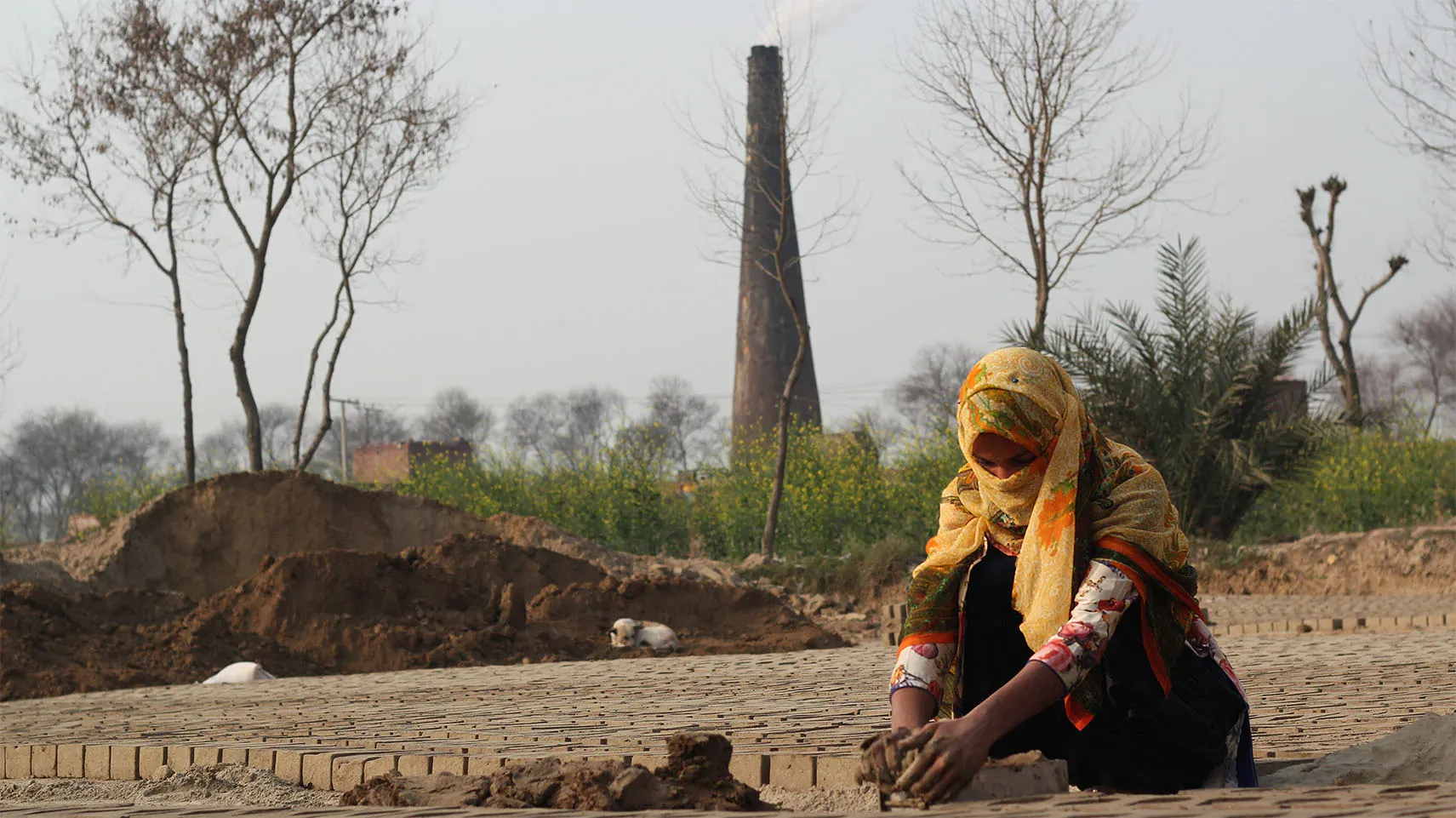 A woman shaping bricks by hand at a kiln, with a smokestack rising in the background